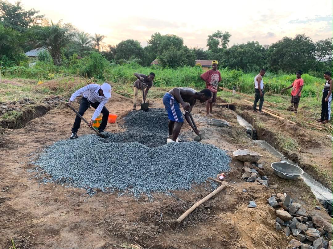 Workers spreading gravel into the foundation trenches of Clinic One at sunset